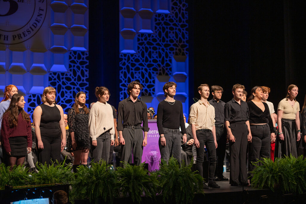 row of students stand on stage behind row of ferns