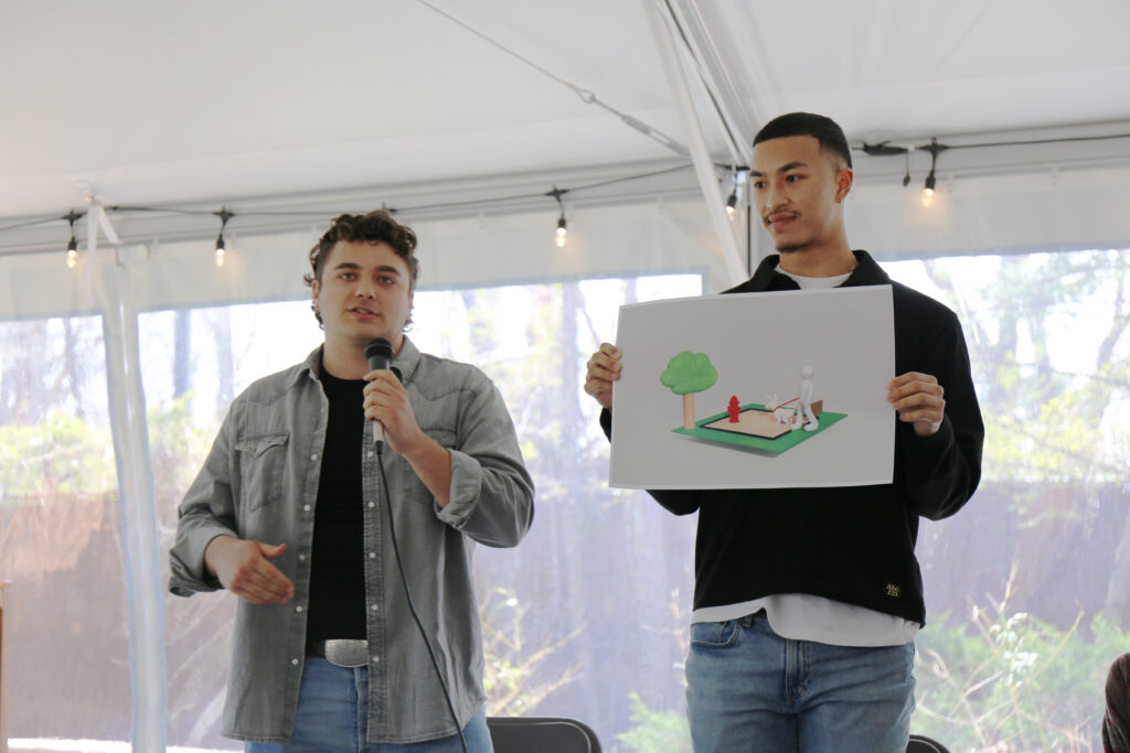 Two people stand while one talks holding a microphone and the other holds up an image on a piece of paper