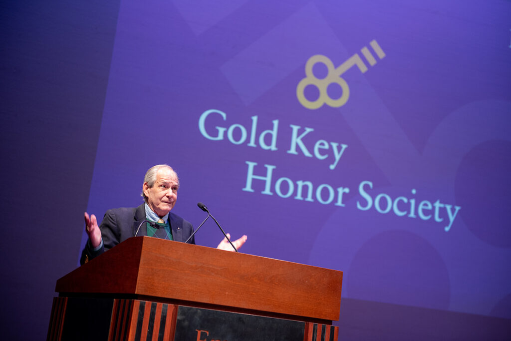 White man with grey hair, wearing a blue and green argyle sweater and navy blazer gesticulates at a podium.