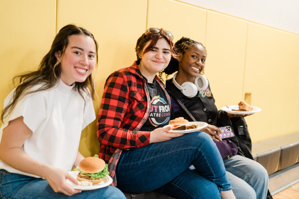 Three people sit while holding plates of food and smiling at the camera