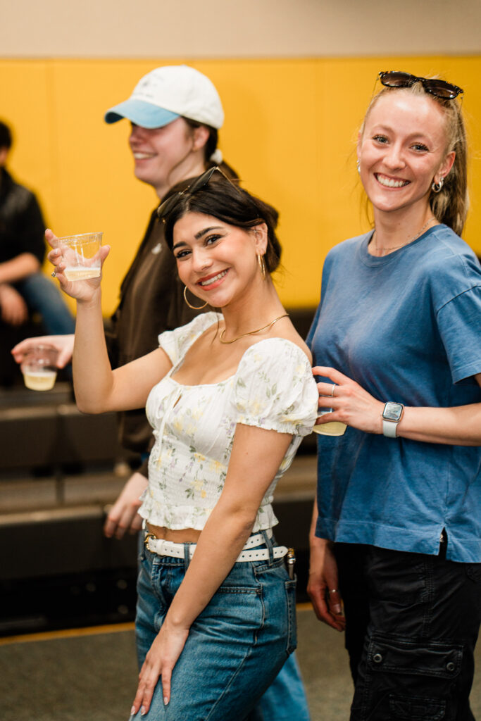Two people smile at the camera while one holds up their drink in a toasting manner