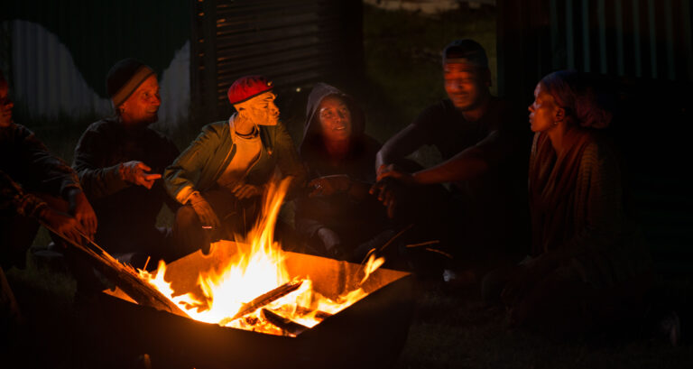 Humans and marionettes, sit around a fire in a crate