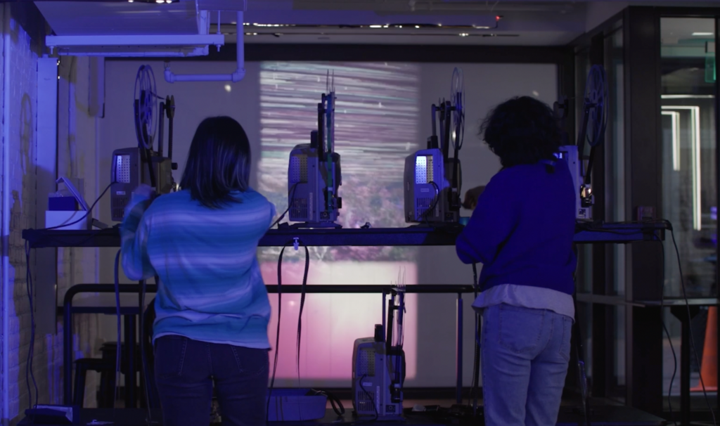 Two people stand behind film projectors.