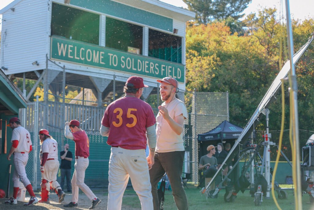 Carson Lund talks to an actor dressed as a baseball player on a baseball field between shots
