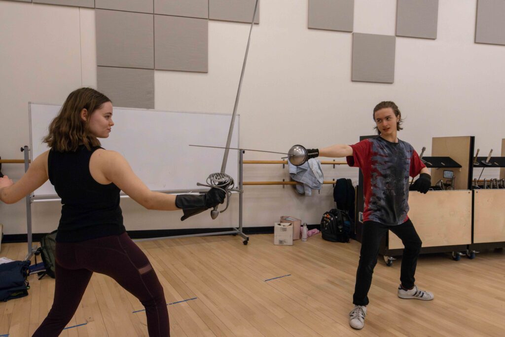 Two people are in fencing position as one of them holds their sword straight out and the other holds their sword upright