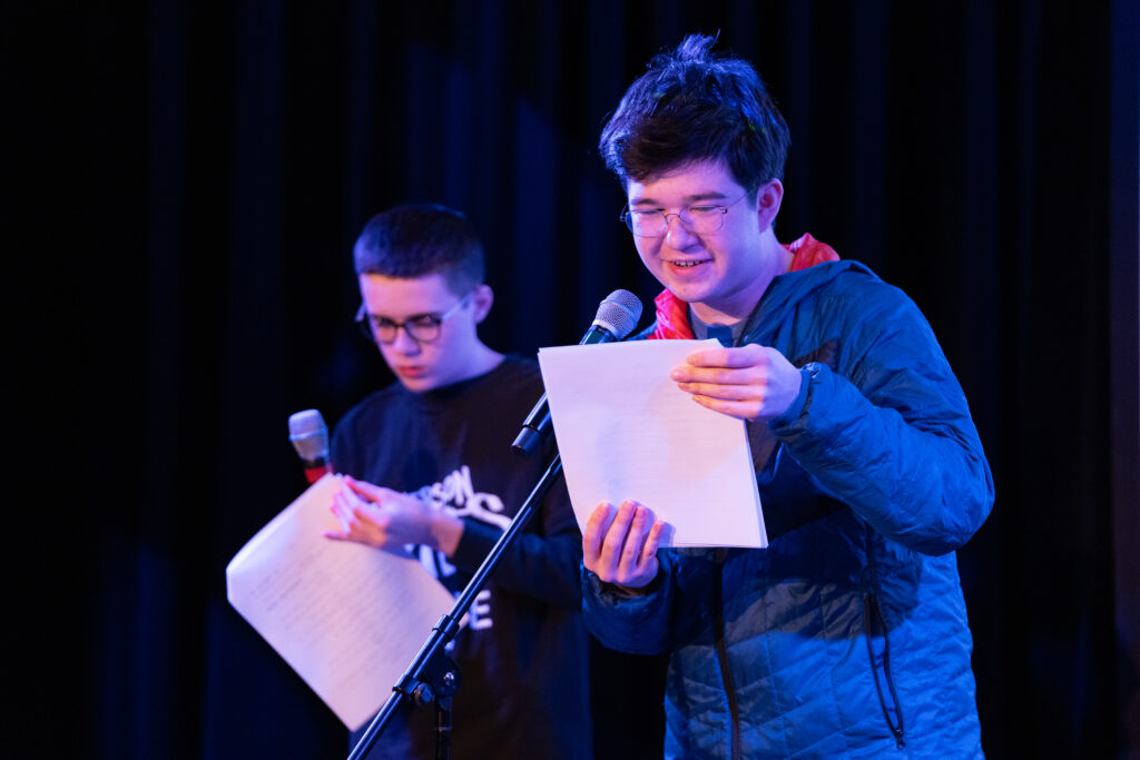 A student uses a microphone to read one of their works during the show while another student is behind them, holding a mic and paper