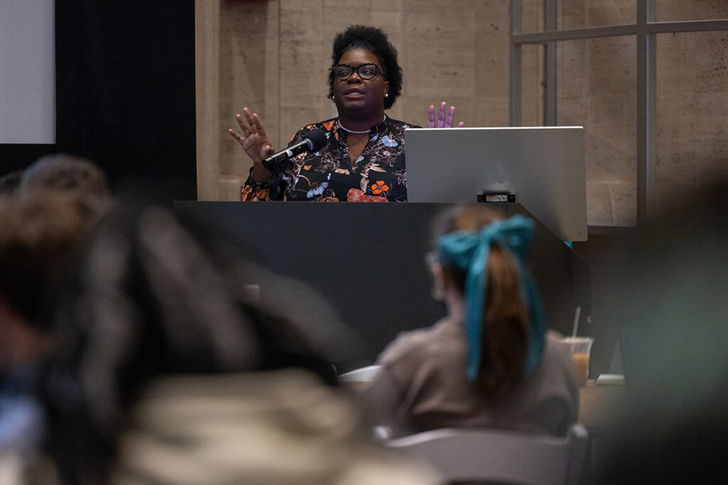 Woman in floral shirt stands behind podium, gesturing