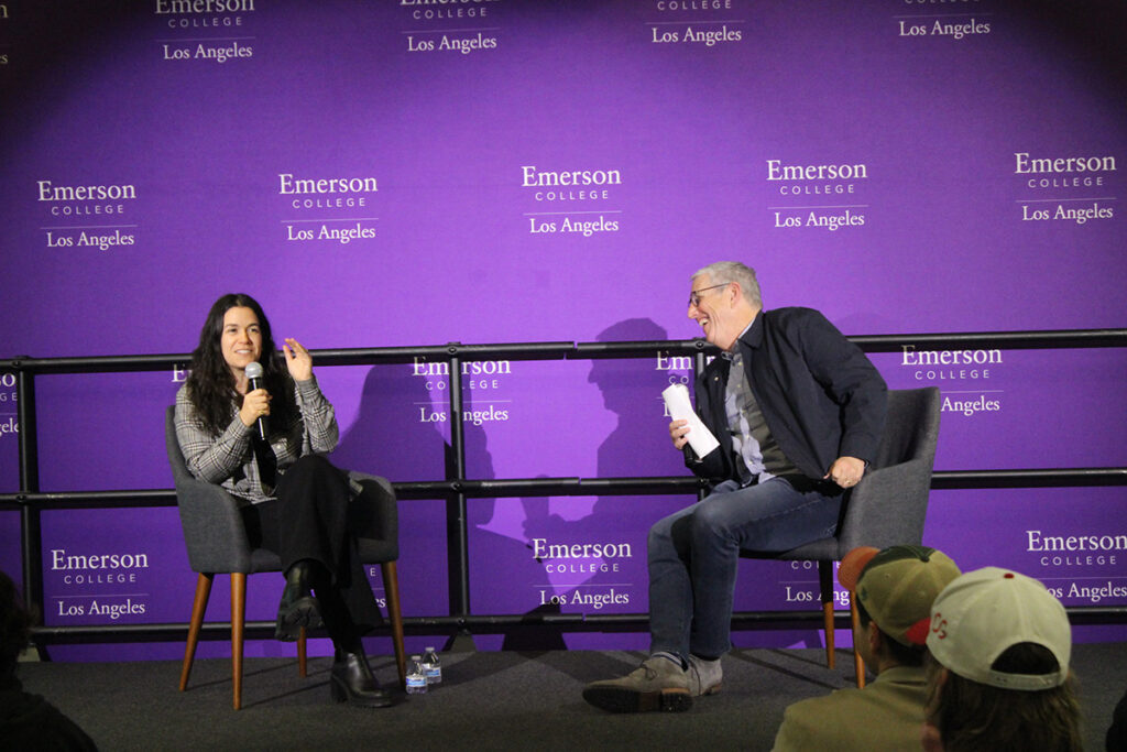 Abbi Jacboson, seated on stage, speaks into mic as Doug Herzog, also seated leans toward her laughing