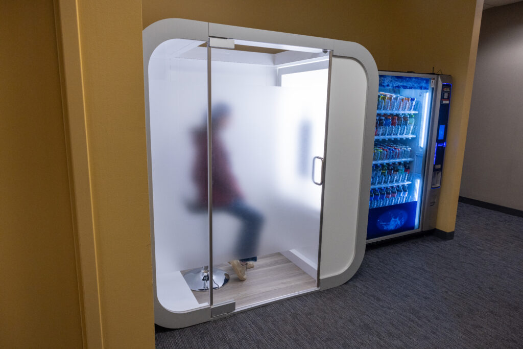 A person sits inside the photo booth with the door closed. The photo booth is next to a vending machine
