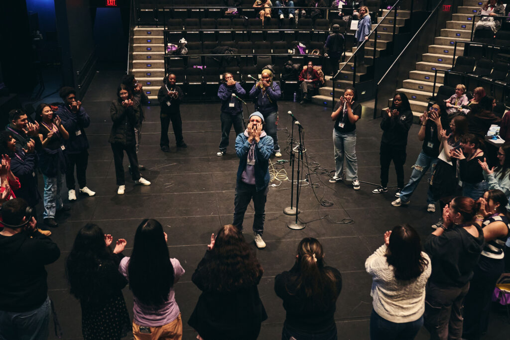One person puts his hands around his mouth leading an exercise on stage while participants are in a circle around them