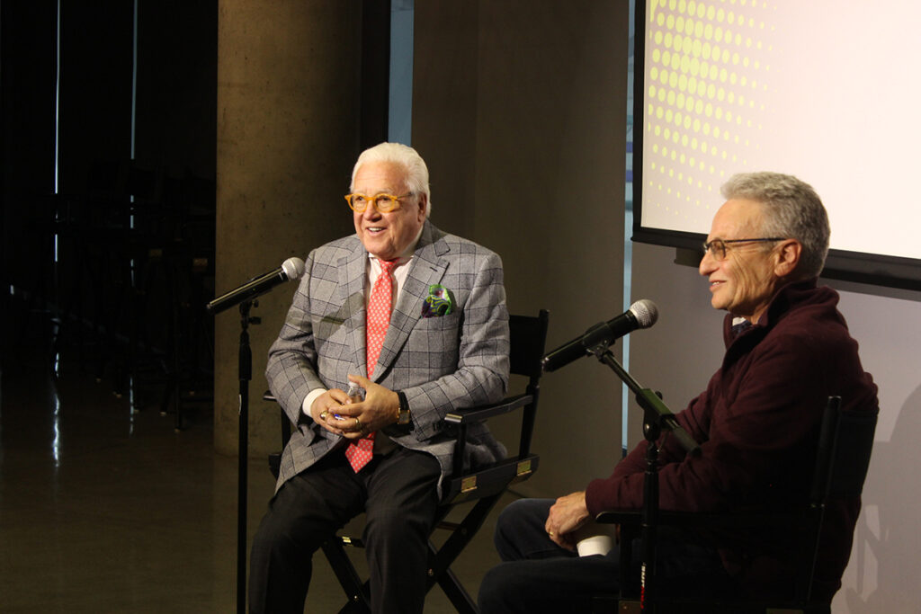 Two men with grey hair, glasses sit and speak into microphones