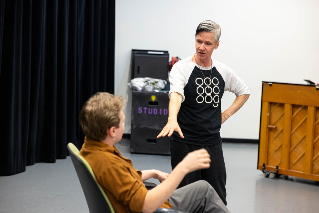 John Cameron Mitchell stands while talking to Asa Dupras is sitting