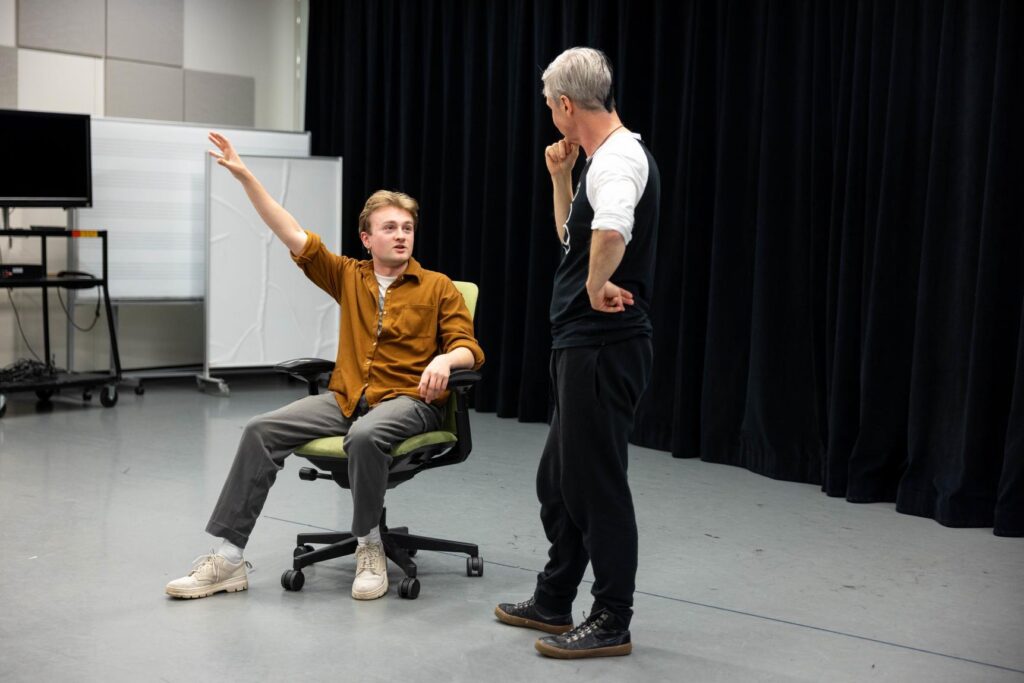Asa Dupras sits in a chair with his arm up talking to John Cameron Mitchell while standing