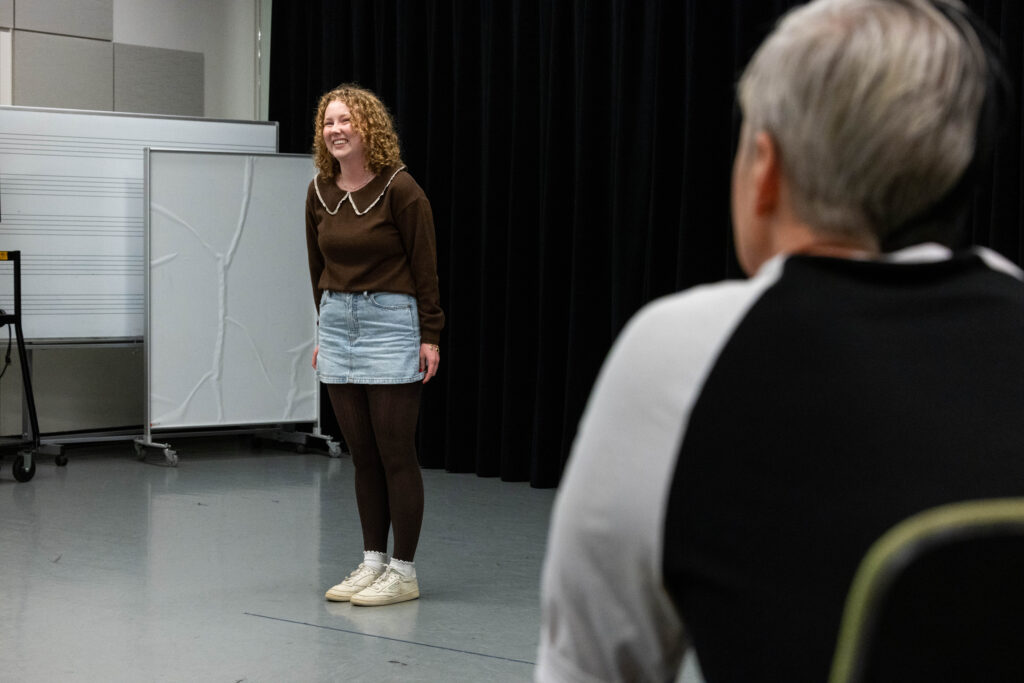 Maggie Cavanaugh smiles with her arms to her side while John Cameron Mitchell looks on