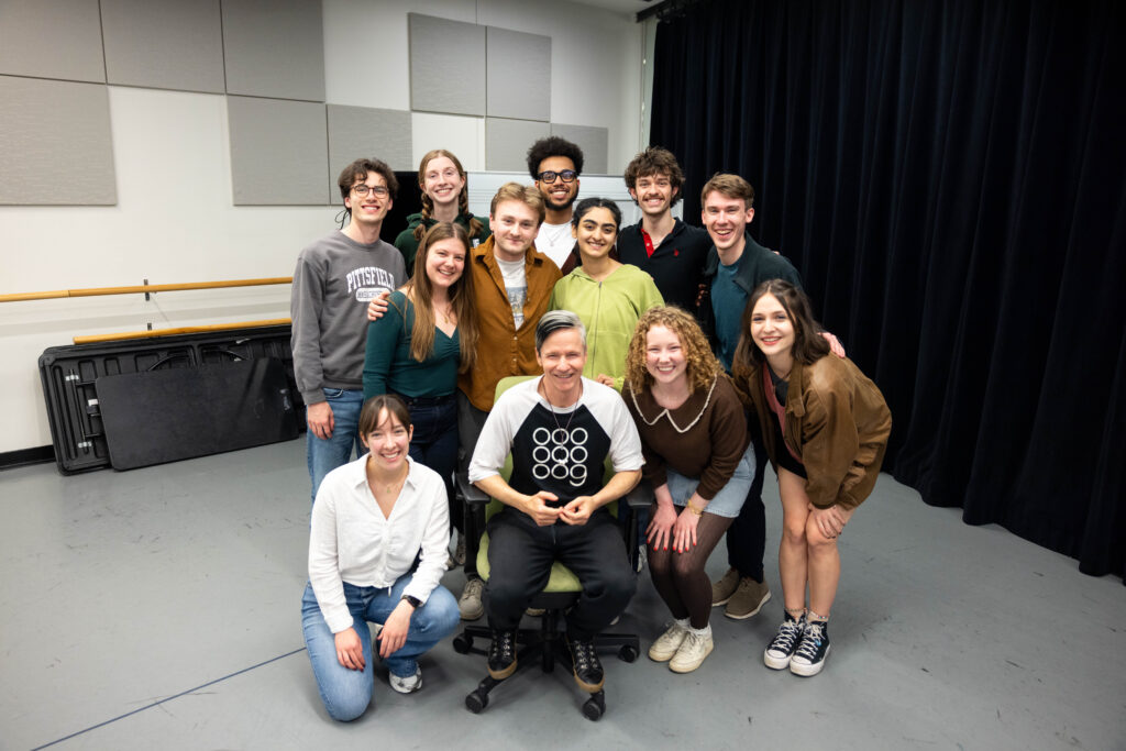John Cameron Mitchell sits in a chair while students stand or crouch near him.