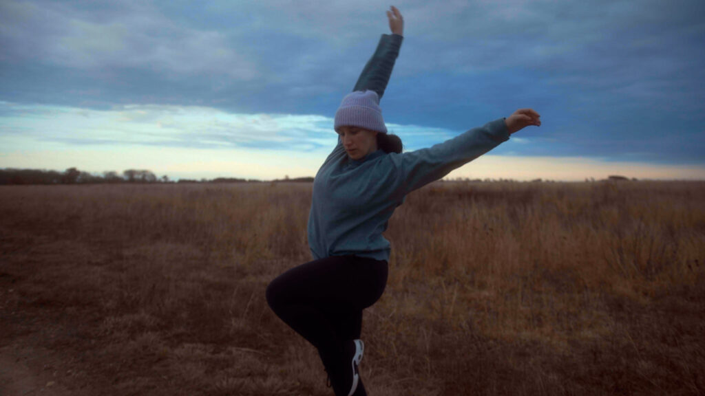 A woman stretch and lifts her leg in a yoga=like pose in the middle of the field with a sunset amid clouds