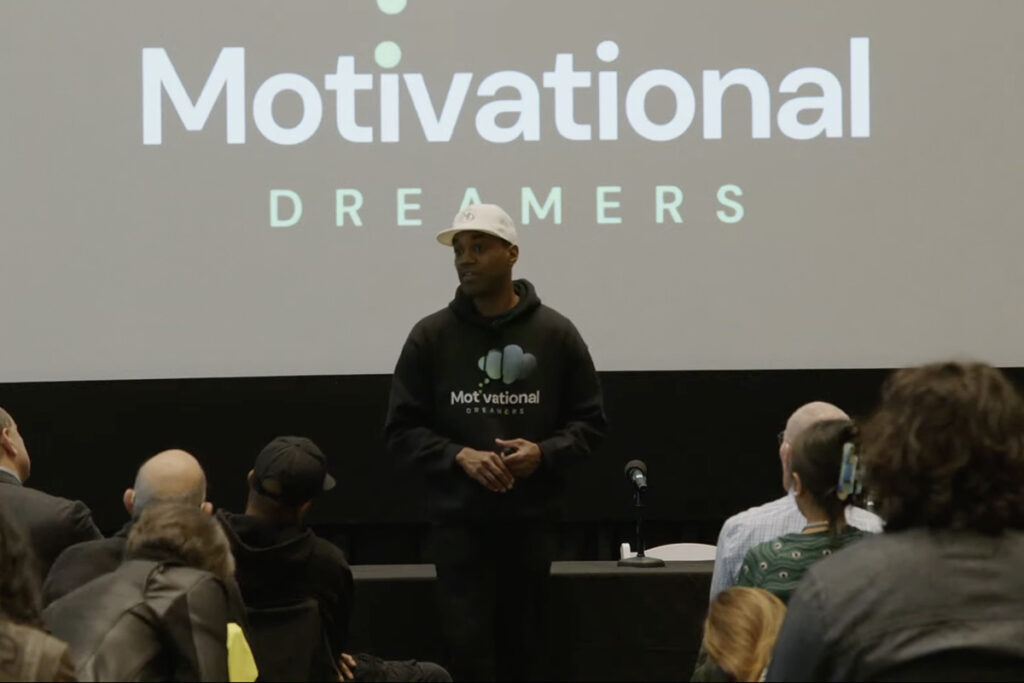 Man in black hoodie with company logo on front, white ball cap, speaks to audience in front of screen reading "motivational dreamers"