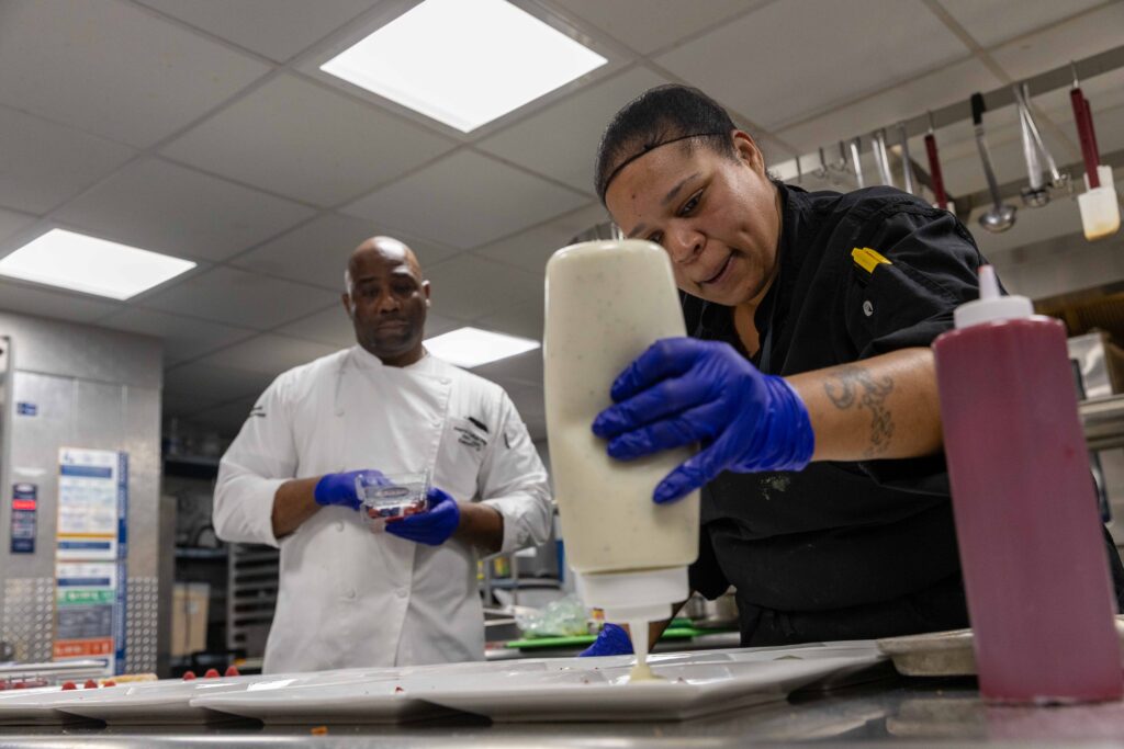 Sus Chef Joanna Cabral squeezes a little bit of something onto a plate while Executive Chef Alex Drumm looks on