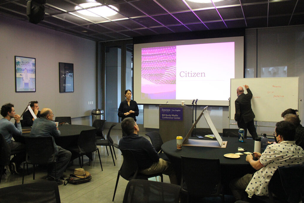 Woman stands in front of screen reading "citizen" as man writes on whiteboard