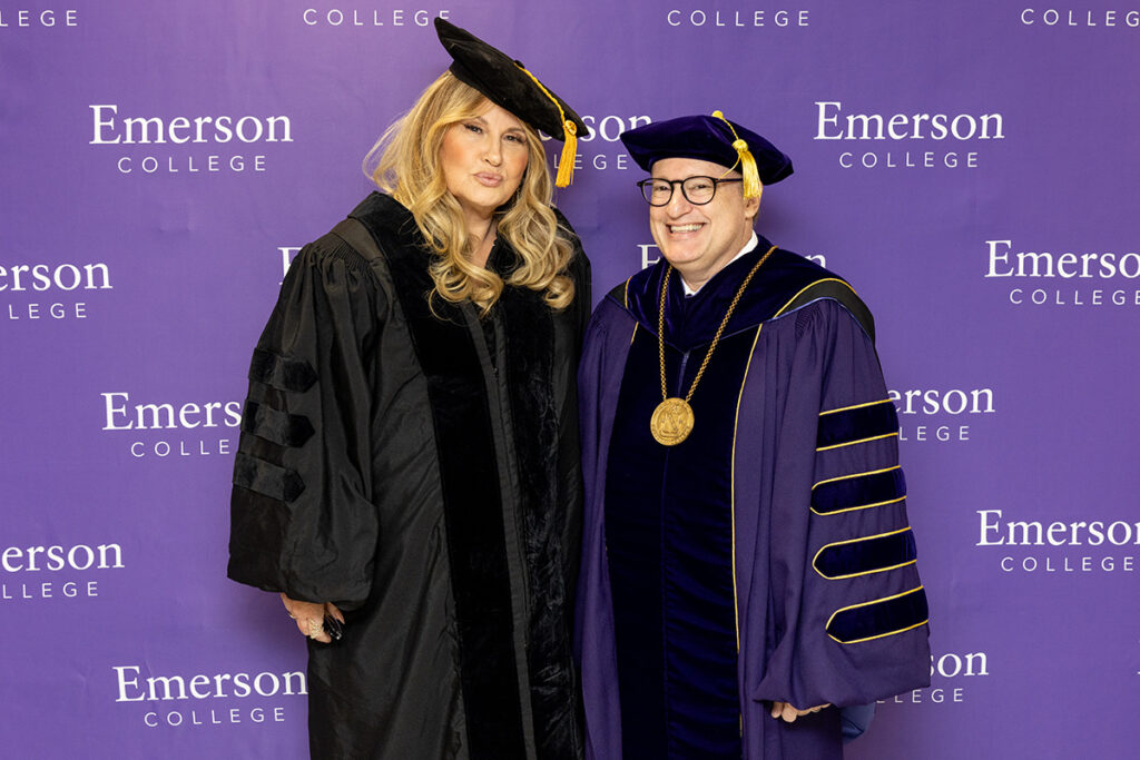 Jennifer Coolidge in cap and gown standing next to Jay Bernhardt in regalia in front of Emerson College step and repeat