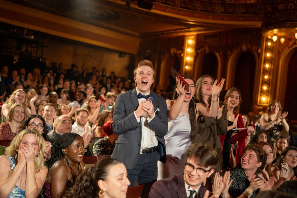 Several people excitedly stand up clapping in the audience