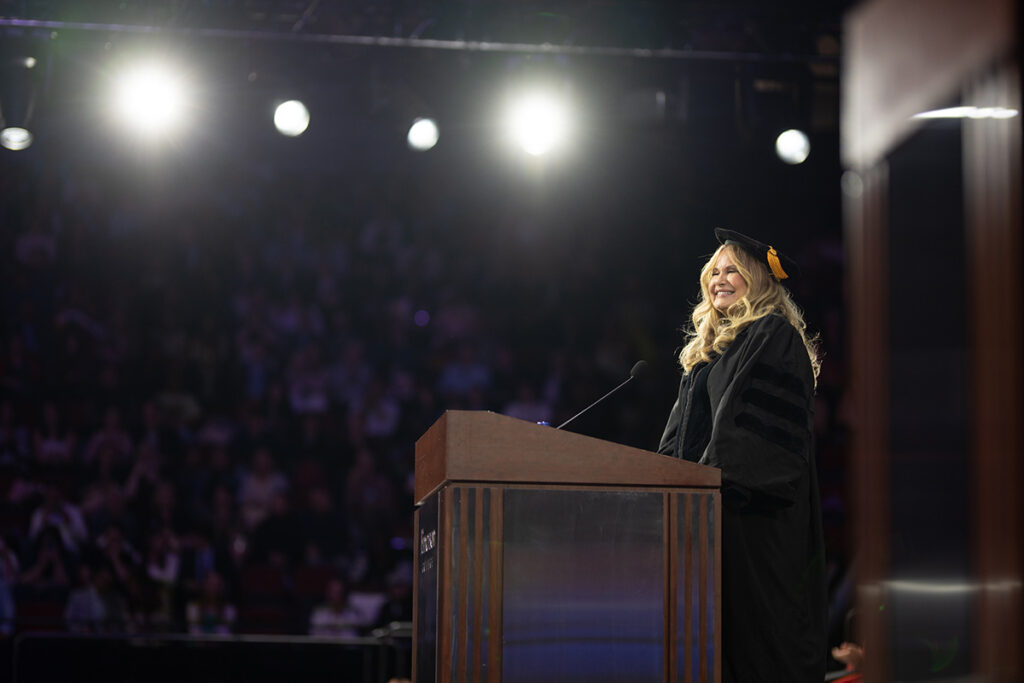 Jennifer Coolidge in cap and gown at podium with spotlights in background