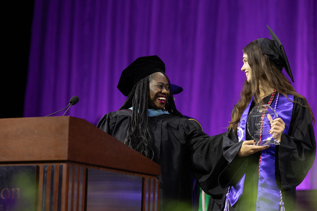 Christie Anglade in black cap and gown gestures to a young woman holding a glass trophy