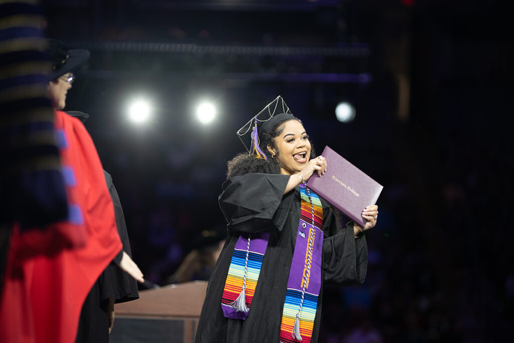 Student in rainbow hood holds purple diploma cover