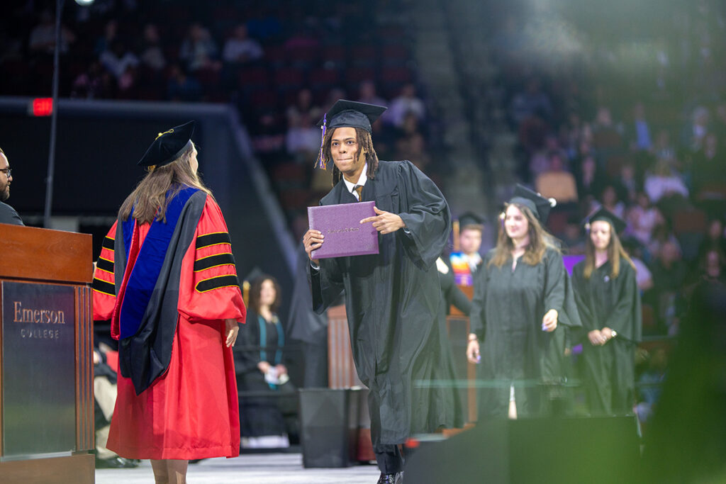 Student in black gown holds purple diploma cover on stage