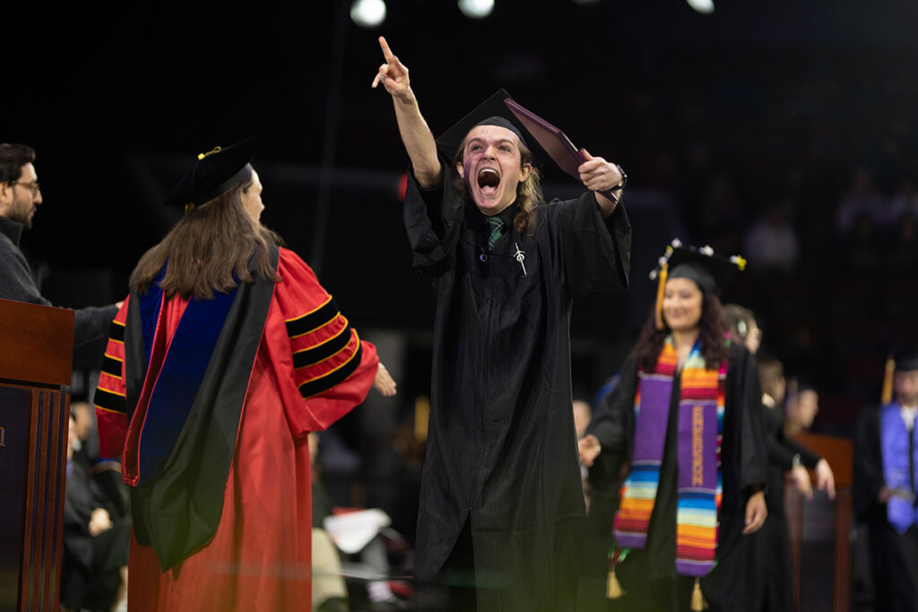 Student in black gown holding diploma cover points into crowd and screams