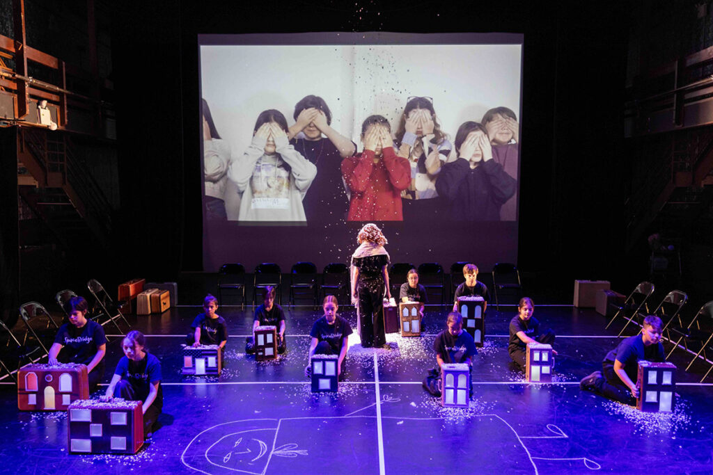Children kneel on stage behind models of buildings as children are projected on screen behind them covering their eyes