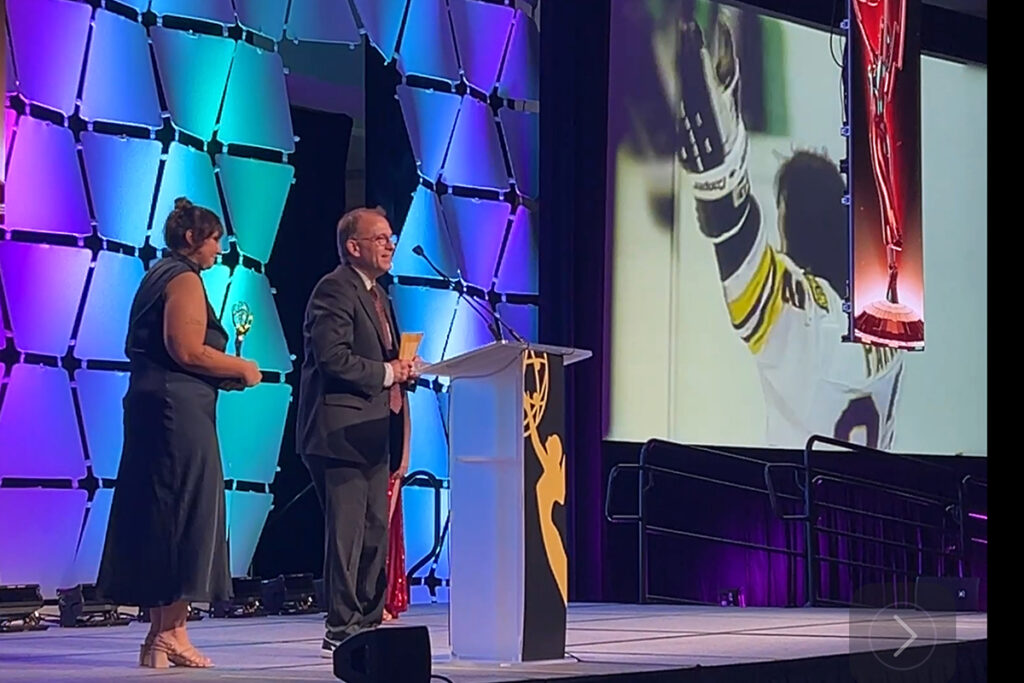 Sean Allen stands at podium as video of a Bruins game is on screen behind him
