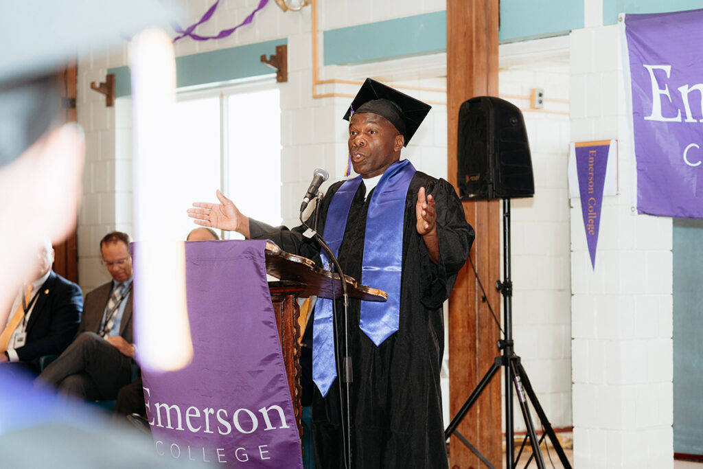 Man in cap and gown gestures behind podium draped in Emerson College banner