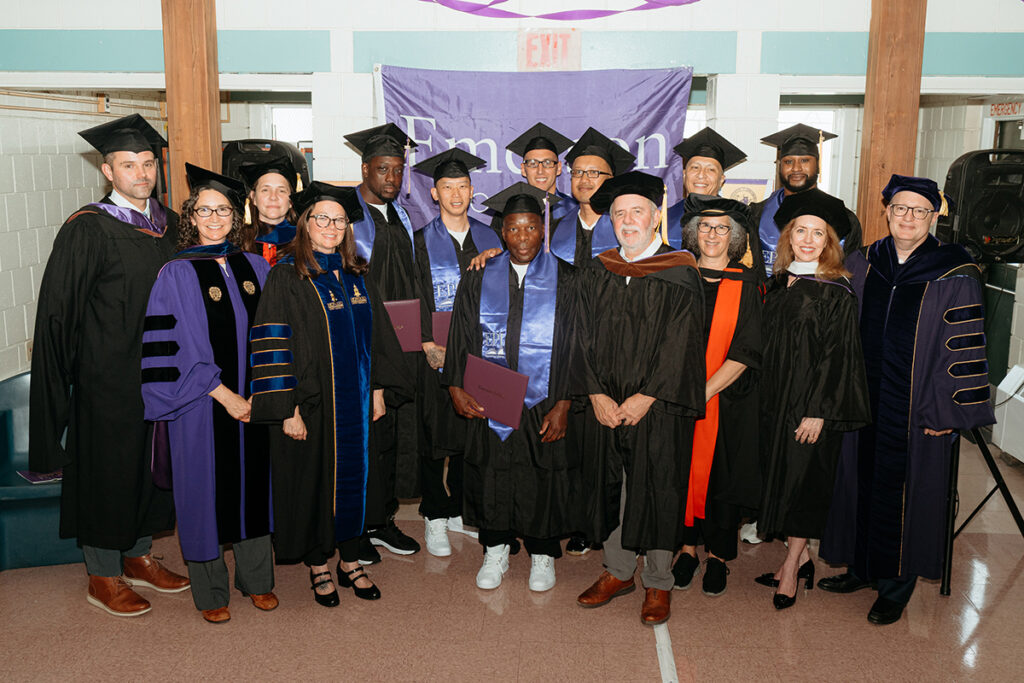 Men and women in academic regalia pose before an Emerson College banner