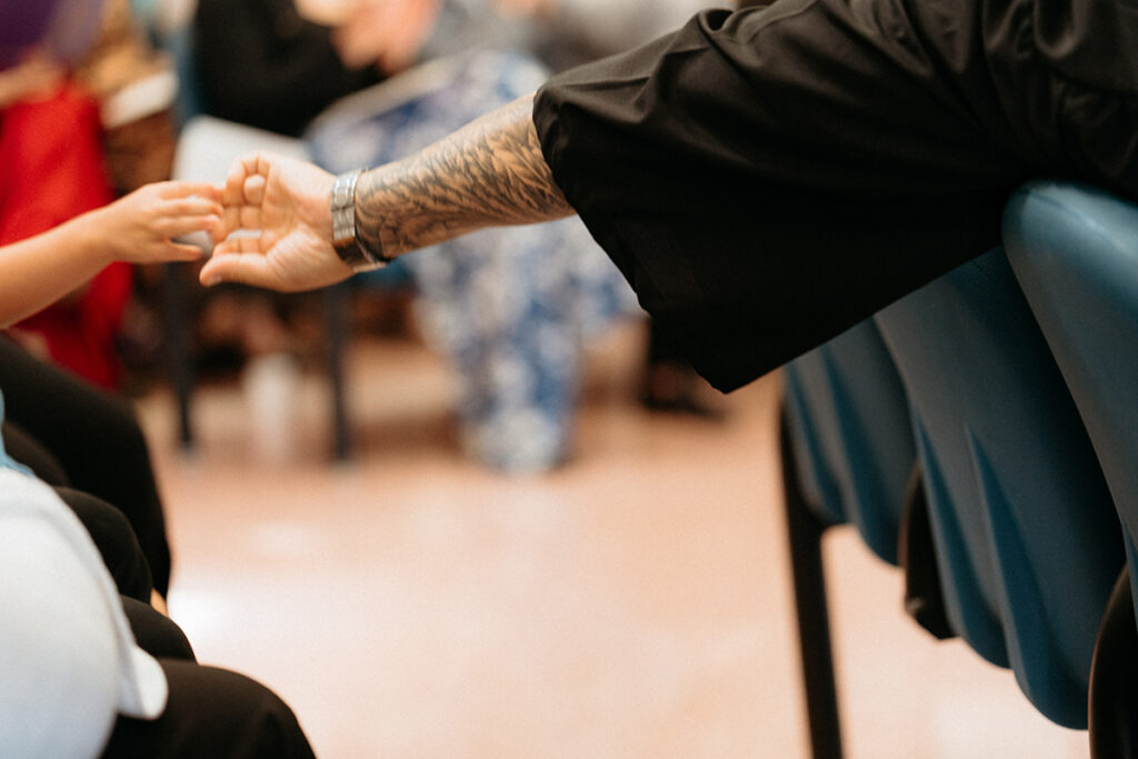 an arm covered in tattoos and an academic gown reaches out to touch the hand of a small child