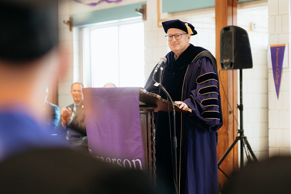 Man in academic regalia stands behind podium with Emerson College banner draped over it