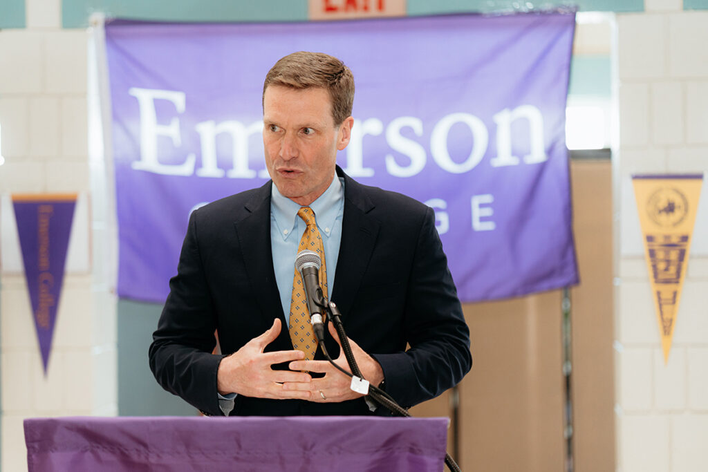 Man in suit and tie speaks behind podium, Emerson College banner behind him