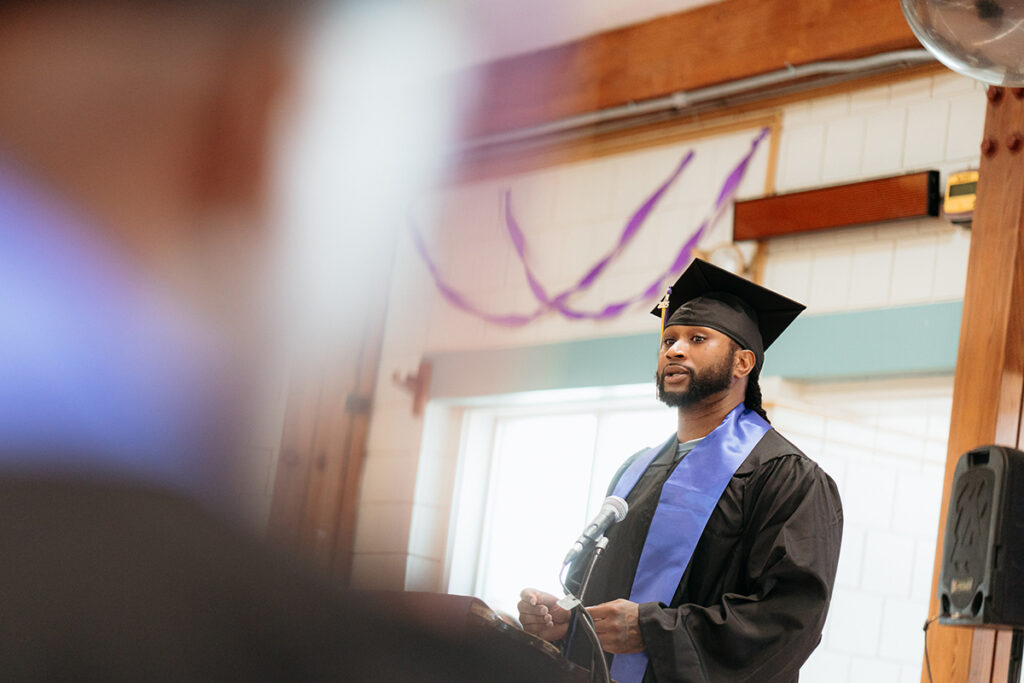 Man in cap and gown speaks at podium in photo shot over the shoulder of another graduate