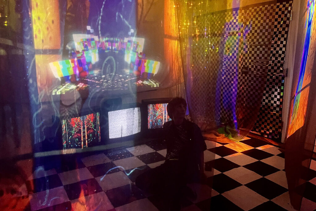 person sits on black and white checkered floor with monitors showing colorful images behind them