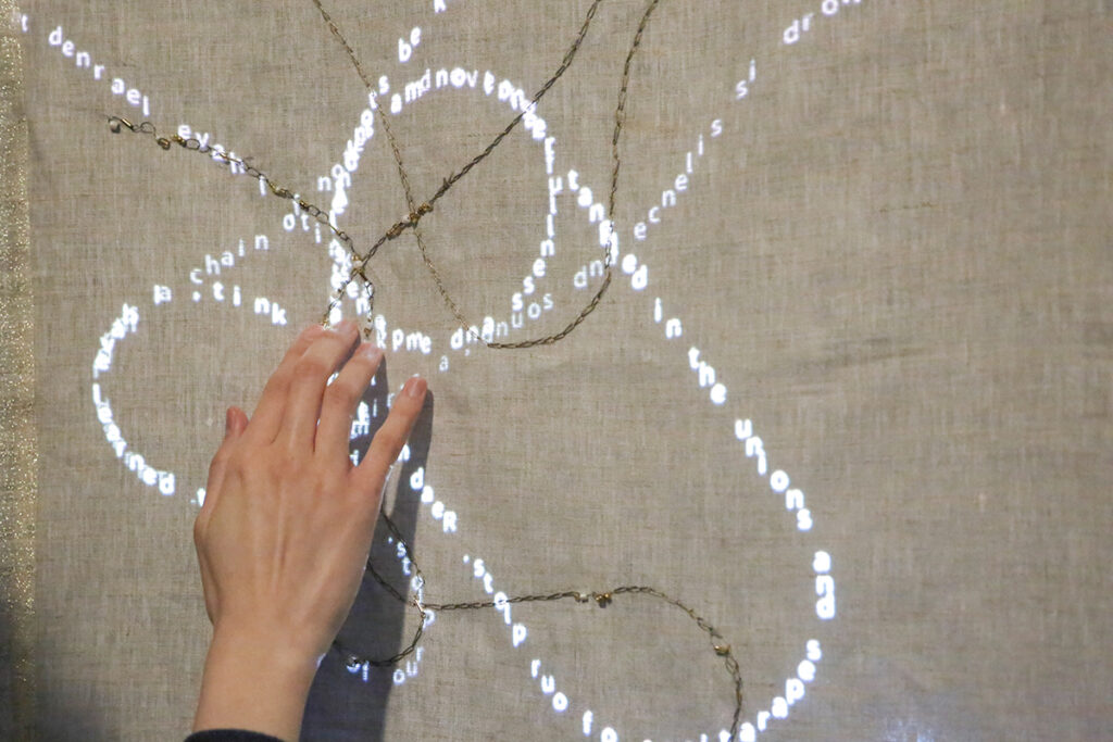 hand reaches up to touch words projected on beige fabric embroidered with gold chain
