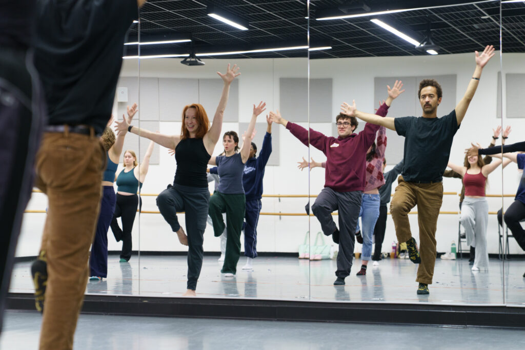 Stephanie Heroux and students look at a mirror wall while spreading their arms wide and having one leg up