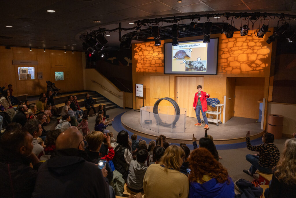 A person in a red coat on stage talks to the audience