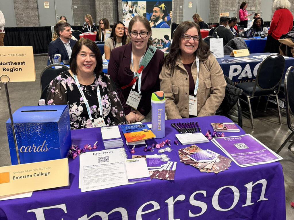 Three women stand behind table festooned with Emerson branded handouts at convention center