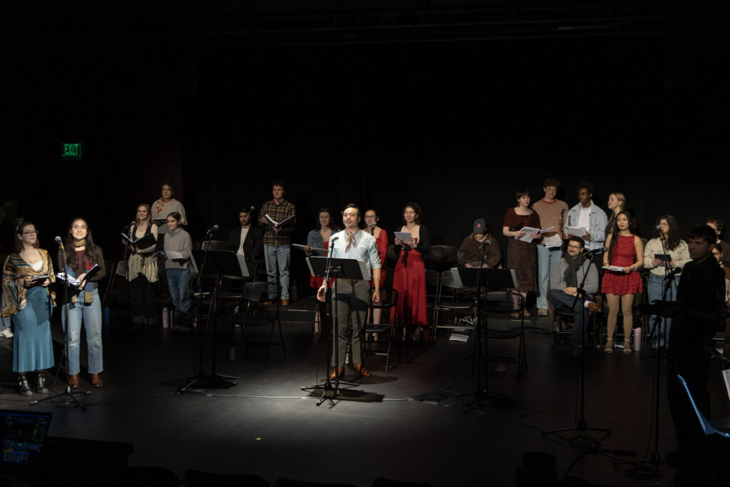 Sam Simakh stands singing with a spotlight on him while the rest of the cast are sitting or standing behind him