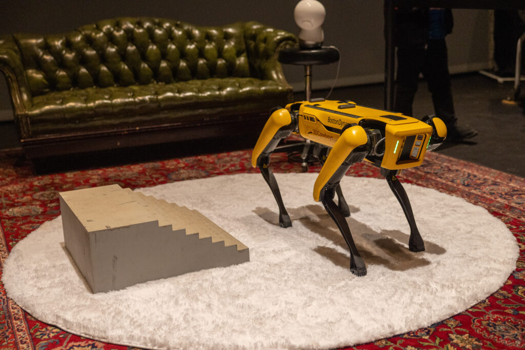 Spot, a robotic dog, stands on a rug with a couch behind it