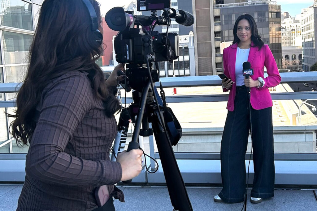 woman in pink blazer reports news on a city rooftop as woman in brown sweater films her