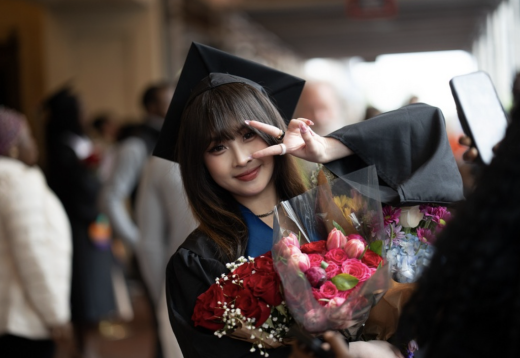 A graduate gives the peace sign while holding flowers