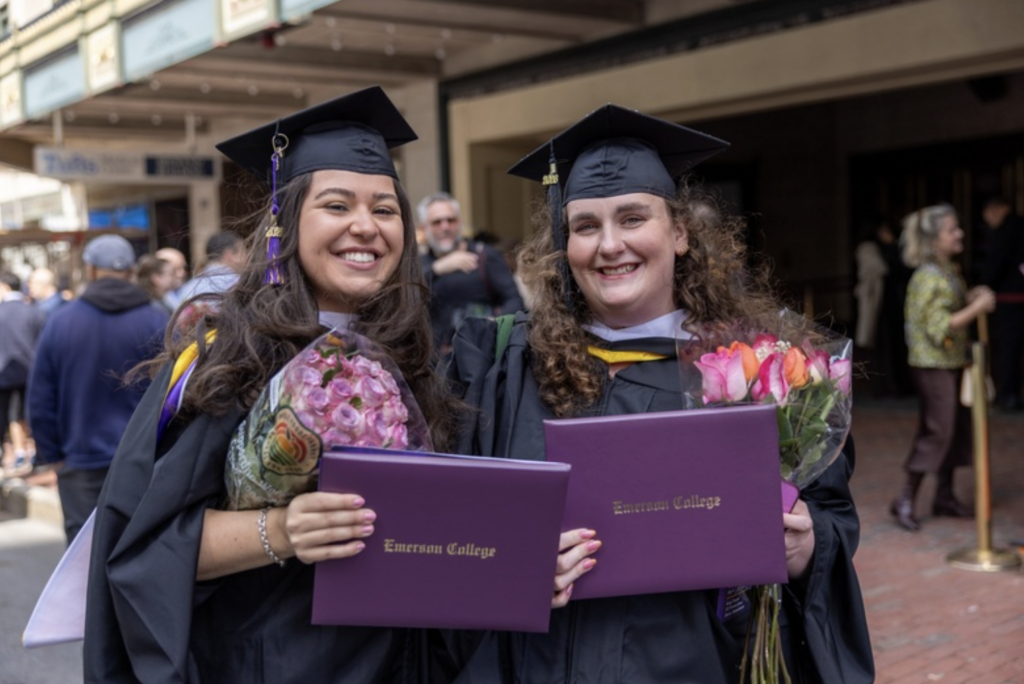 Two graduates smile while holding their diplomas