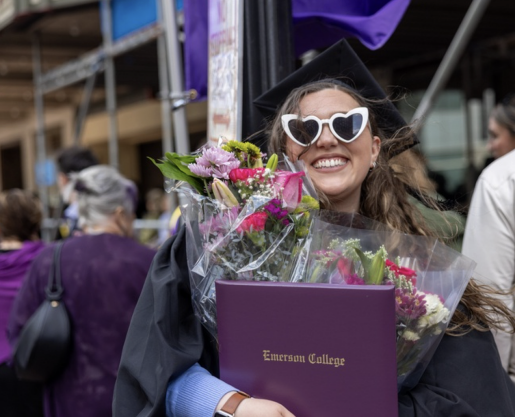 A graduate with heart-shaped sunglasses smiles while holding flowers and their diploma