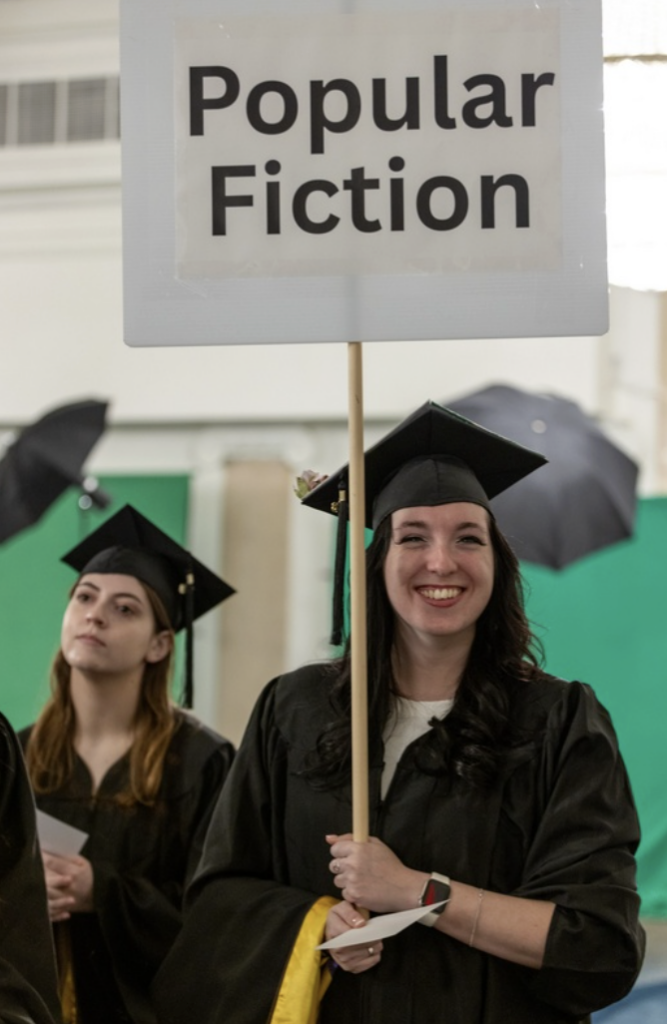 A graduate smiles while holding a sign that says Pop Fiction